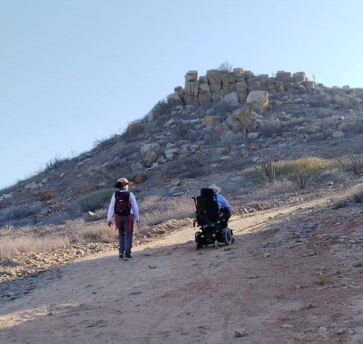 Una persona en silla de ruedas junta a una persona caminando subiendo un sendero en un cerro.