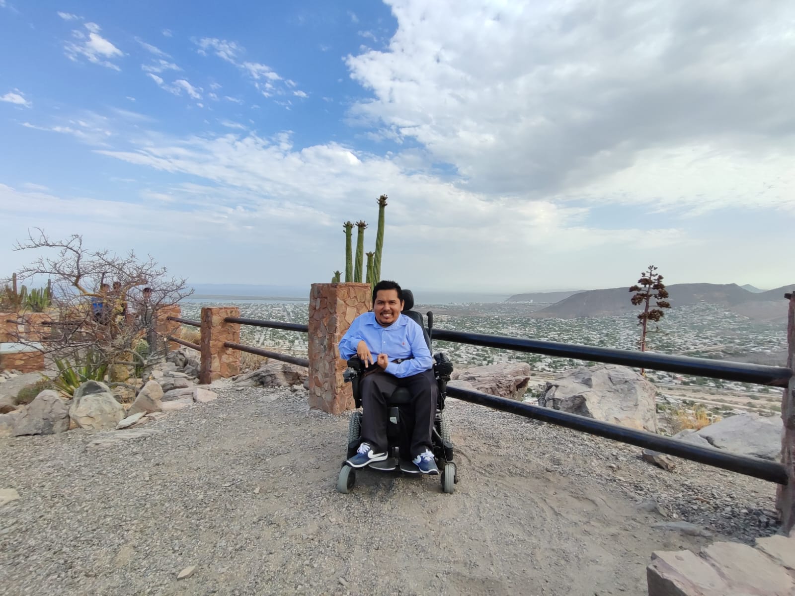 Una persona en silla de ruedas en un mirador en un cerro con algunos cactus y la vista de la ciudad al fondo.