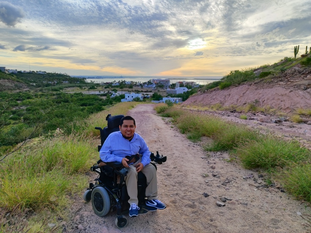 Una persona en silla de ruedas en un camino en un cerro, el cielo y parte de una bahía de fondo