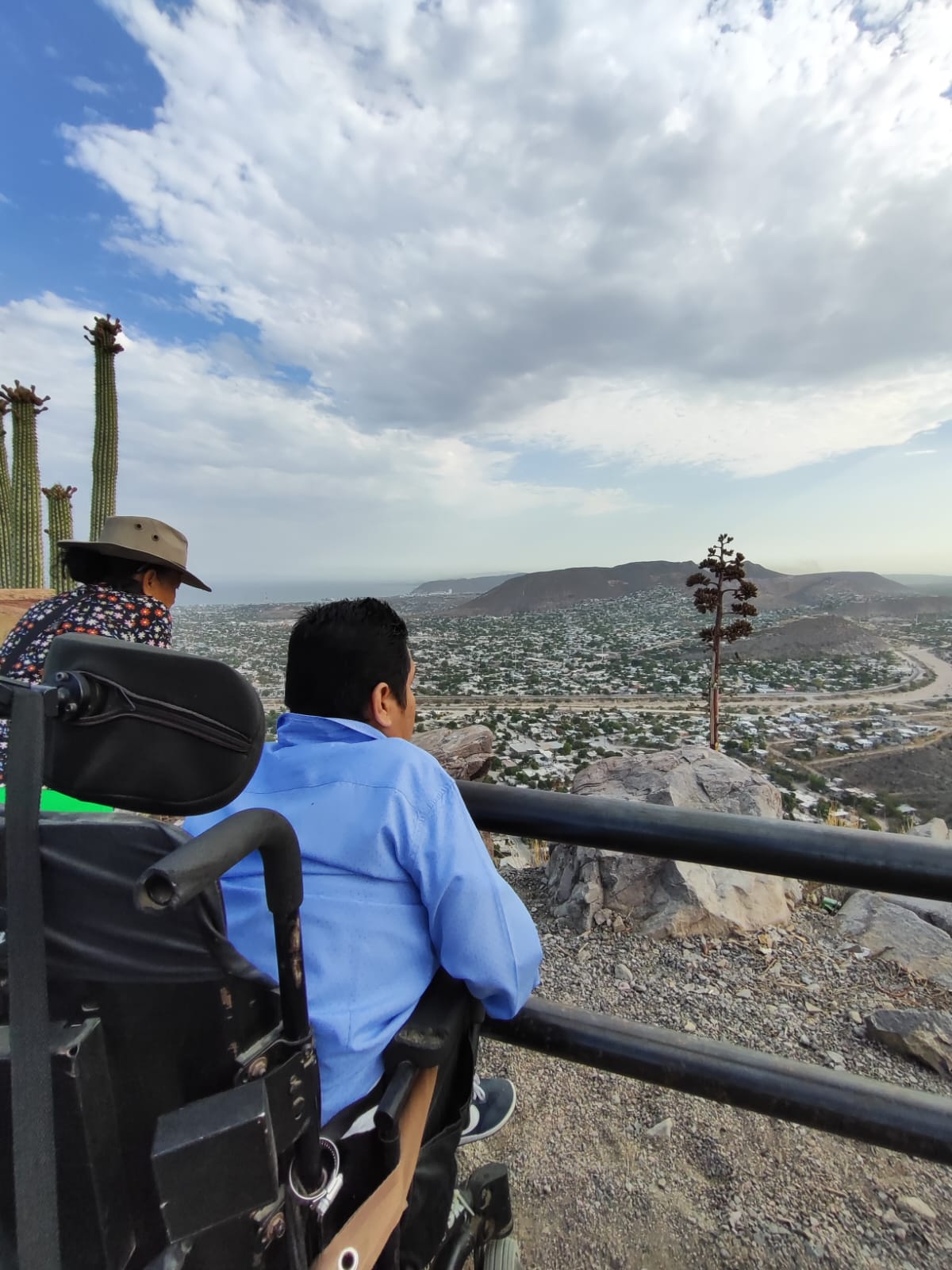 Una persona en silla de ruedas junto a otra persona de pie en un mirador viendo hacia el horizonte.