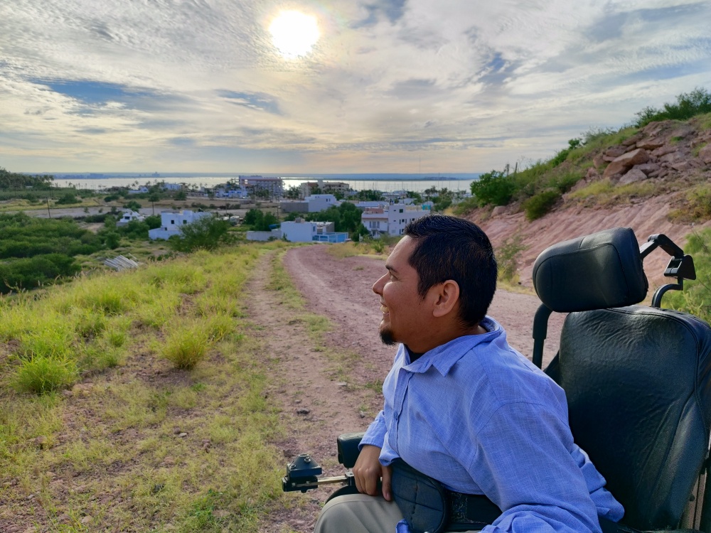 Una persona en silla de ruedas en un camino de tierra con el atardecer y el malecón de fondo